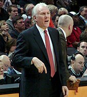 A man, wearing a black suit, white shirt and red tie, is standing in front of the spectators.