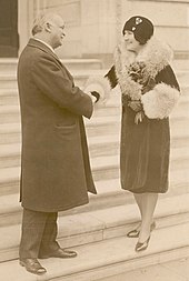 Photograph of a man (left) and woman (right) shaking hands on an outdoor staircase. He is wearing a long wool coat and she is wearing a velvet coat with fur on the cuffs and collar, and holding a bouquet of roses in her free hand.