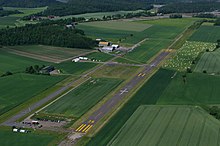 A short, asphalted runway runs though the middle, surrounded by green fields. To the left is a taxiway, and at the top of the runway is a small terminal building