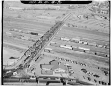 An aerial view of a rail yard, in which a number of freight train cars are visible, predominantly boxcars. A road bridge crosses over the rail yard and connects to a highway in the background.