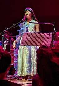Ichiko Aoba stands on stage during a performance, dressed in a light-colored dress with ruffled details. She is singing into a microphone, with a music stand holding sheet music in front of her. The background includes other musicians and instruments.