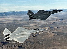 The two YF-23 production models flying over a desert, the first one named "Gray Ghost" painted in charcoal gray is at the left of the second one named "Spider", painted light gray, which is in the immediate front