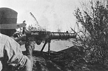 A soldier lies among the grass in the prone position behind a machine gun which he is holding in the shoulder.