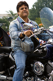 A man with black hair, wearing jeans and a denin jacket, is sitting atop a black motorcycle.