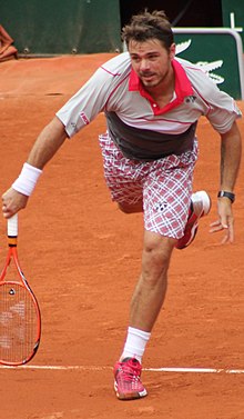 A man wearing a light gray shirt with a red collar, a red plaid-designed shorts, and red shoes finishes his tennis service motion as he brings his red racket down in front of him