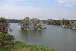 A small round island with trees in the middle of a large body of water, with more trees on the shore on either side