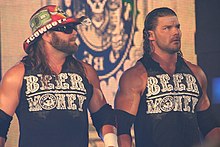 Two adult white males wearing black shirts with "Beer Money" in green text on the front. Both have long black hair, one wearing a cowboy hat.