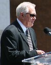 Rod Philips wearing black sunglasses talking to a crowd on a podium with a microphone