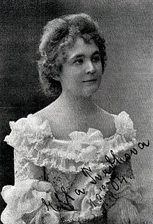A white woman in a ruffled white dress with a wide neckline. Her hair is in a bouffant updo.