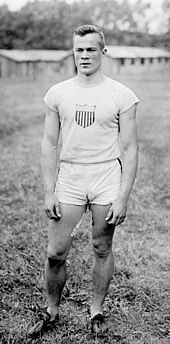 A young man wearing an all-white singlet and shorts stands in a grassy field.