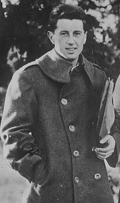 A black-haired man in a dark trench coat poses the picture, looking towards the camera