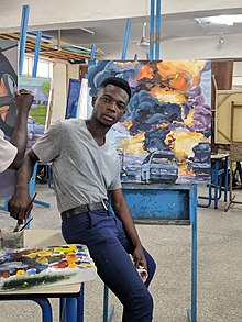 Oscar Korbla Mawuli Awuku resting on a table, holding a paintbrush while looking at camera