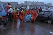 Scene of first responders wearing orange uniforms carrying light blue bags away from the disaster scene. A crowd with umbrellas surround the disaster workers and several cars are parked. The day is cloudy.