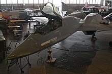 A YF-23 undergoing restoration at the US Air Force Museum, one other plane can be seen at top left, another at top right; jeeps, machines, benches, cables and a plaque can be seen