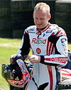 A man in his late 30s is wearing white motorcycling overalls with red stripes looking at his left hand and holding a crash helmet in his right hand