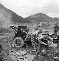 Soldiers firing a howitzer against a mountainous backdrop.