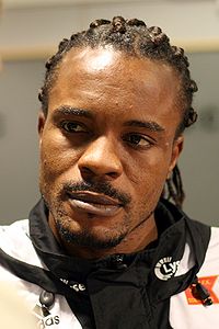 A photograph of a black man wearing a white and black training jacket. The man is looking away from the camera.