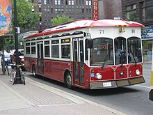 a bus adjacent to a large sidewalk in an urban area, it is red on the underside and white on the upper half, with a red roof. it seems slightly dated in design style, and has two doors on one side.