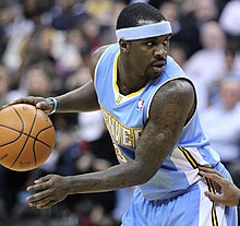 A basketball player in a light blue jersey wearing a light blue headband while dribbling a basketball.