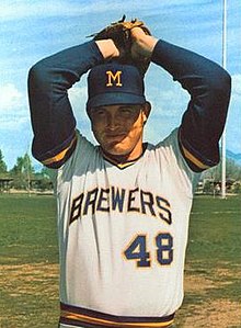 A man wearing a white Brewers jersey with yellow and blue trim at the neck and sleeve openings and a blue cap with an "M" on the front holding his hands in his glove above his head