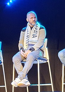 Man sitting in chair with a scarf over his shoulders and a microphone in his hand