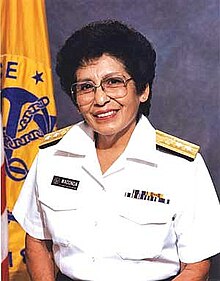 A short-haired woman in glasses in a white uniform in front of a flag