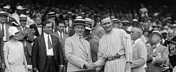 Two men stand in front of a crowd shaking hands. The man on the left of the photo is wearing a tan suit and hat and the man on the right is wearing a light-colored pinstriped baseball uniform.