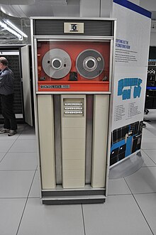 A large cabinet, about the size of an upright refrigerator, with a glass-covered top part holding two reels of magnetic tape, and a bottom part with control buttons framed by vertical channels.