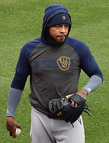 A man in a navy blue and gray hoodie and gray pants holding a baseball on a green field