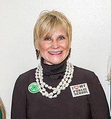 A white woman with blonde hair is wearing a brown turtleneck and a large white necklace; she is looking and smiling into the camera.