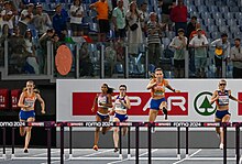 Photo with a frontal view of Femke Bol jumping over a hurdle and Cathelijn Peeters, Lina Nielsen, Louise Maraval, and Line Kloster running between hurdles while being watched by spectators behind a fence