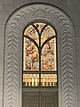 A stained glass window with branches depicting cherry blossoms and leaves, surrounded by gold. The window is surrounded by carved masonry, depicting branches and cherry blossoms stretching from the top of the window to the bottom along an arch.