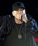 A man in a black cap, t-shirt and jacket, and dog tag performing with a microphone in his hand