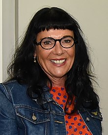 A smiling woman looking into the camera, with brown eyes, long dark hair and fringe, wearing round black glasses and a denim jacket over a spotted orange blouse.