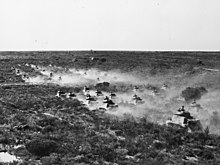 Several columns of tanks advance across an arid landscape, churning up clouds of dust