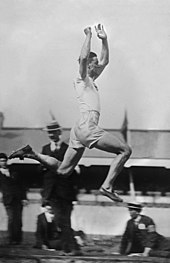A young man wearing an all-white singlet and shorts is in mid-air while competing in the long jump competition at the 1920 Summer Olympics.