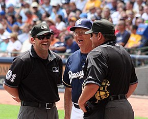 A man in a navy blue baseball jersey standing on a field between two men in black umpiring outfits