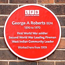 Circular red plaque, with six lines of text reading "George A Roberts BEM / 1890 to 1970 / First World War Soldier / Second World War Leading Fireman / West Indian Community Leader / Worked here from 1939". Above those, the text and logo "London Fire Brigade".