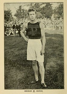 white man with short hair looking directly at the camera, one hand on hip and the other arm hanging straight down, wearing baggy white athletic shorts and a dark sleeveless jersey with a large "P" on the chest