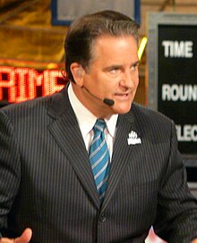 Candid photograph of Mariucci seated behind a desk on a TV set wearing a dark pin-striped suit, blue striped tie and a headset
