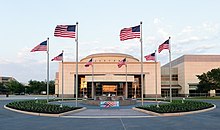 Above the doorway of a large, relatively plain rectangular structure with a short dome are the words "George Bush Library." In front of the building is a circular courtyard with a water fountain; eight American flags are positioned evenly around the circle.