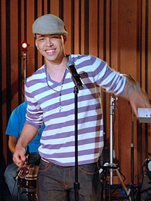 A man looking straight, slightly inclined to the right, wearing a beret, purple shirt with white stripes, a rosary and a pair of jeans, with a microphone in front of him.