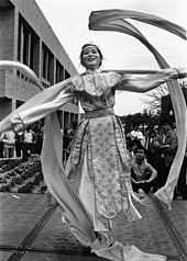 Woman in a long dress dancing and smiling with spectators and campus buildings in the background