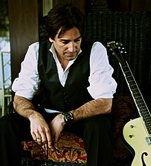 Country singer Steve Azar, wearing a white shirt, vest, and pants, leaning against a chair, with an electric guitar propped up next to him
