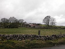 Green farmland on an overcast day, with a stone wall in the foreground, and somewhat dilapidated stone buildings bordered by trees in the background