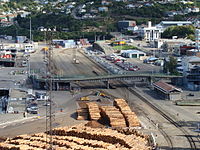 Lyttelton railway station yard. Features of interest include the Oxford Street overbridge (middle), Lyttelton station building (right, in front of the bridge), shore-end of wharves 2 and 3 (left, opposite the end of the bridge), Lyttelton rail tunnel portal (obscured, centre background), and Lyttelton road tunnel portal (centre-right background). Also shown are two of the ports main sources of traffic: timber and motor vehicles.