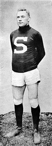 A young man stands at attention in a rugby uniform: a dark colored sweater emblazoned with a large white 'S', white shorts, and dark knee-length socks.