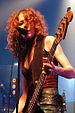Melissa Auf der Maur—a Caucasian woman with long red hair wearing a red top and leather skirt—plays bass guitar and sings into a microphone