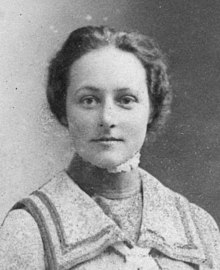 Head and shoulders photograph of a woman facing the camera, shoulders slightly turned, her hair up, with high collar undergarment and a square neck blouse. Her stare is very direct.
