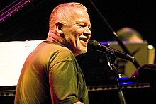 An image of Joe Sample smiling while sitting at a piano with a microphone attached.